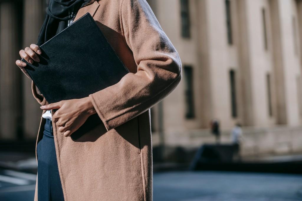 A professional in a camel coat and black hijab holding a large black folder in front of a classical government or courthouse building